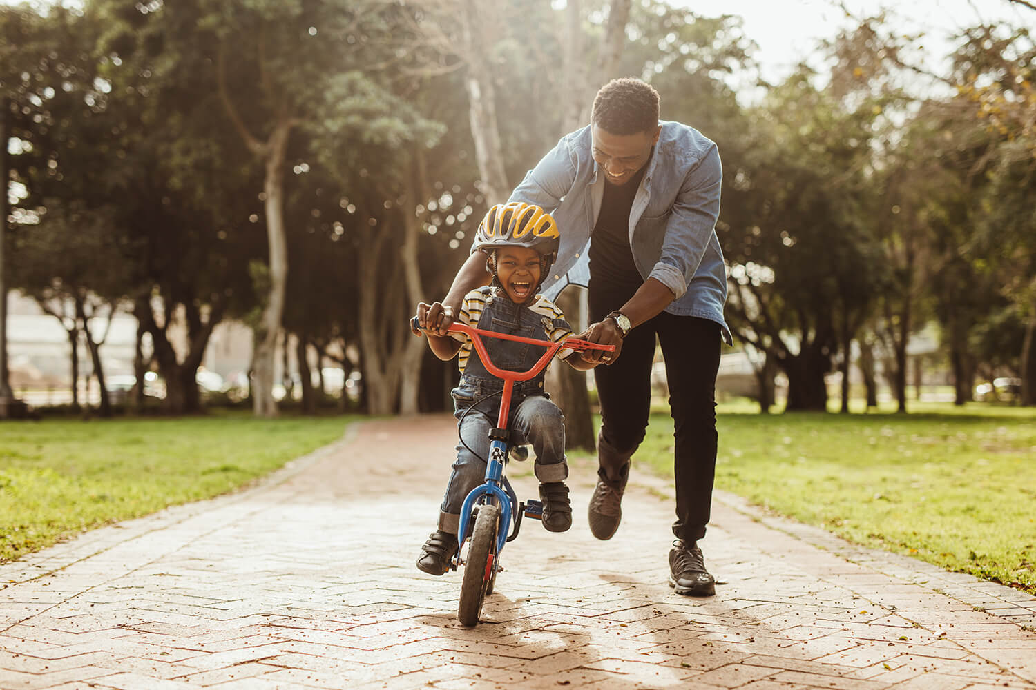 A smiling father helps his young son learn to ride a bike in a Macclesfield park on a sunny day.