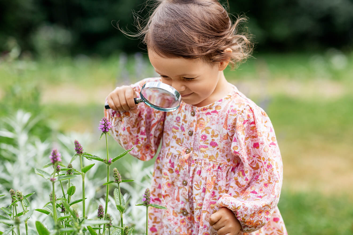 A young girl in a pink dress looks at flowers using a magnifying glass.